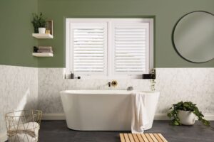 White louvred shutters fitted above a bath in a green and marble bathroom